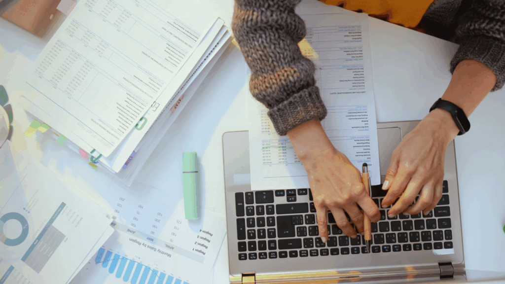 View from above of a table filled with papers, notebooks, a highlighter, and a laptop