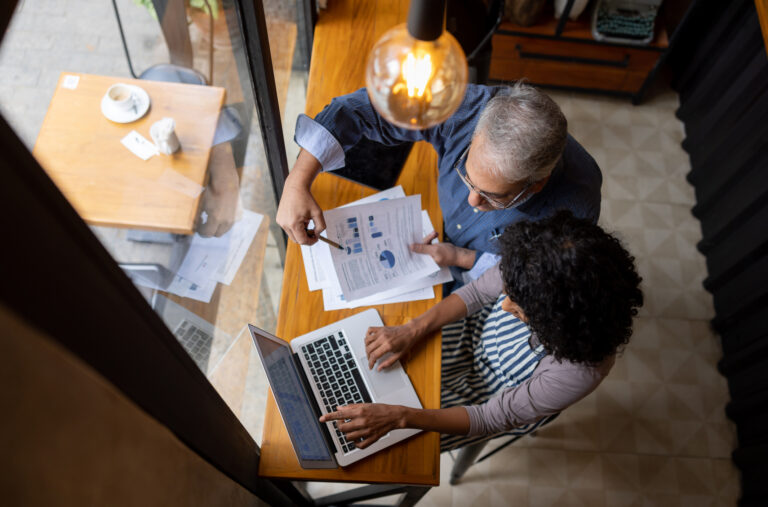 A man and woman sit at a table reviewing papers and a laptop together