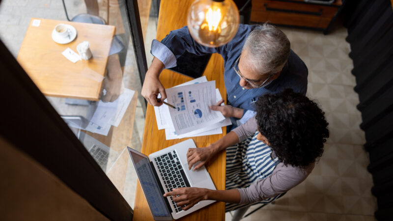 A man and woman sit at a table reviewing papers and a laptop together